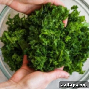 Hands gently massaging fresh kale leaves with olive oil and salt in a clear glass bowl, a vital step for creating a tender and palatable kale salad.
