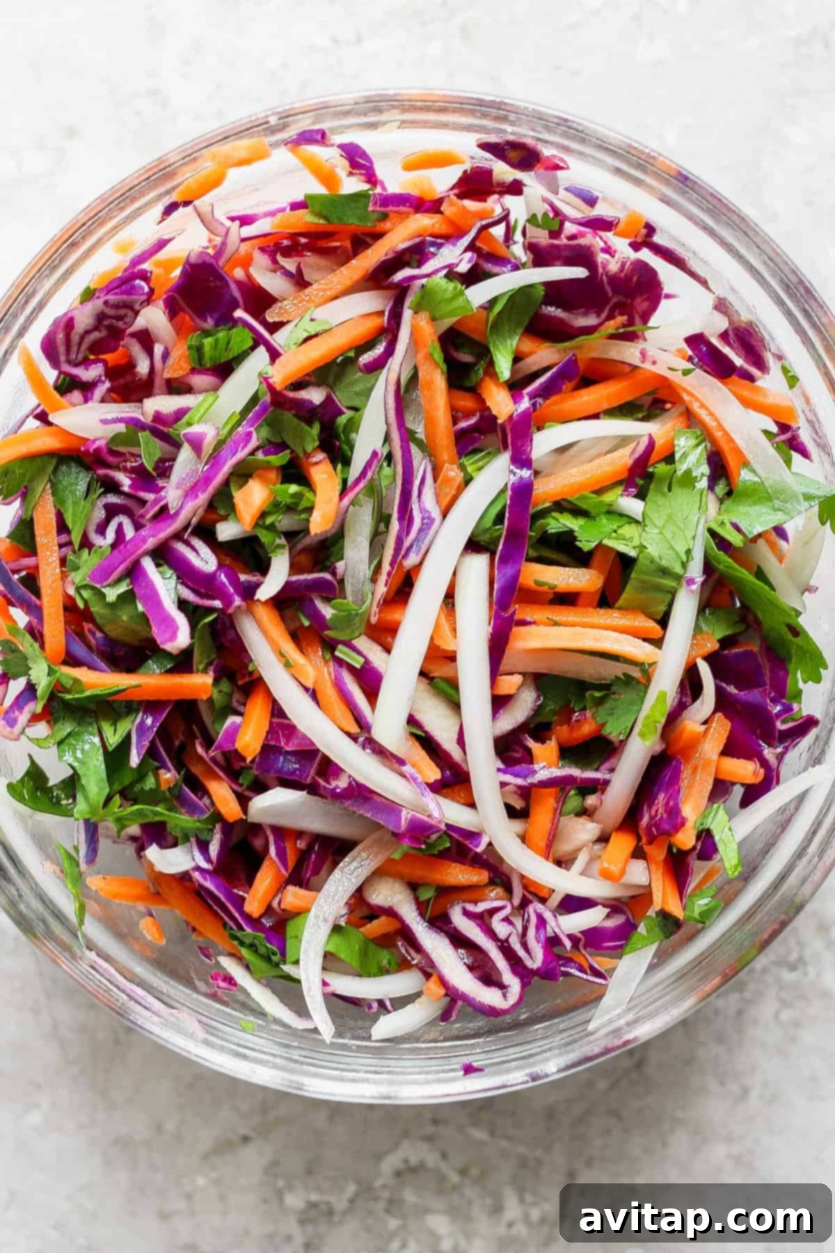 A vibrant, colorful cabbage salad featuring shredded purple cabbage, carrots, white onion, and cilantro, dressed with a tangy vinaigrette, in a clear bowl on a white background, ready for assembly into wraps.