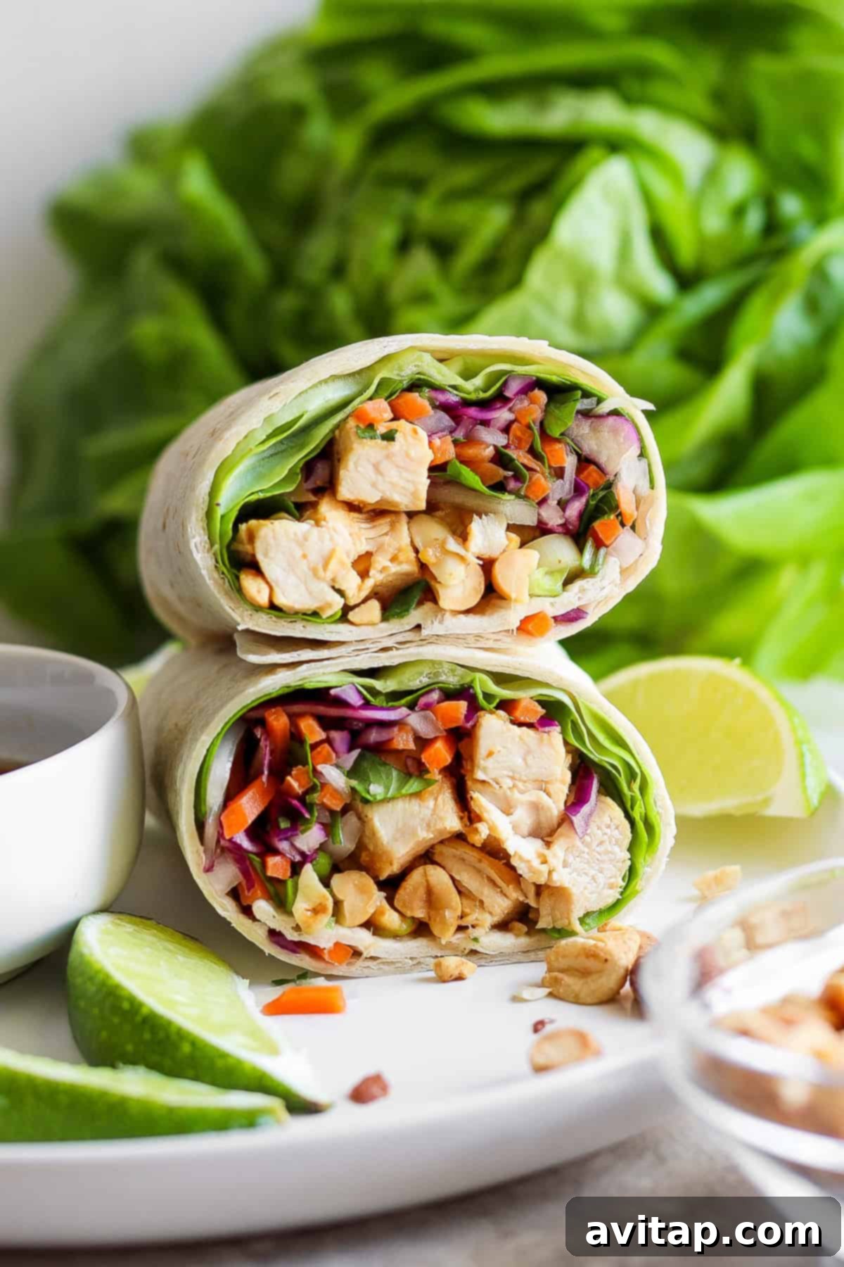A beautifully presented Peanut Chicken Wrap, sliced in half, revealing a colorful interior of chicken, purple cabbage, carrots, and peanuts, served on a white plate with lime wedges and a side of peanut dipping sauce. Fresh butter lettuce is visible in the background.