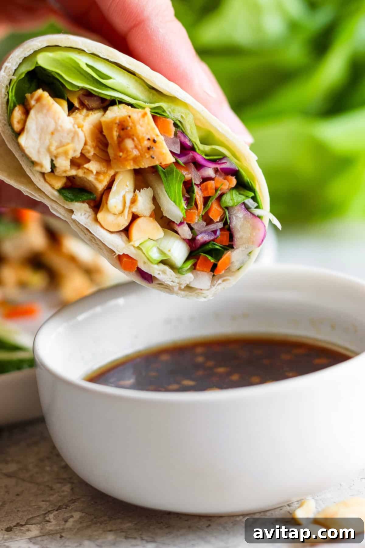 A hand holding a fresh, vibrant spring roll or wrap, filled with tender chicken, colorful vegetables, and a sprinkle of cashews, positioned next to a small bowl of creamy dipping sauce, highlighting the deliciousness and ease of eating.