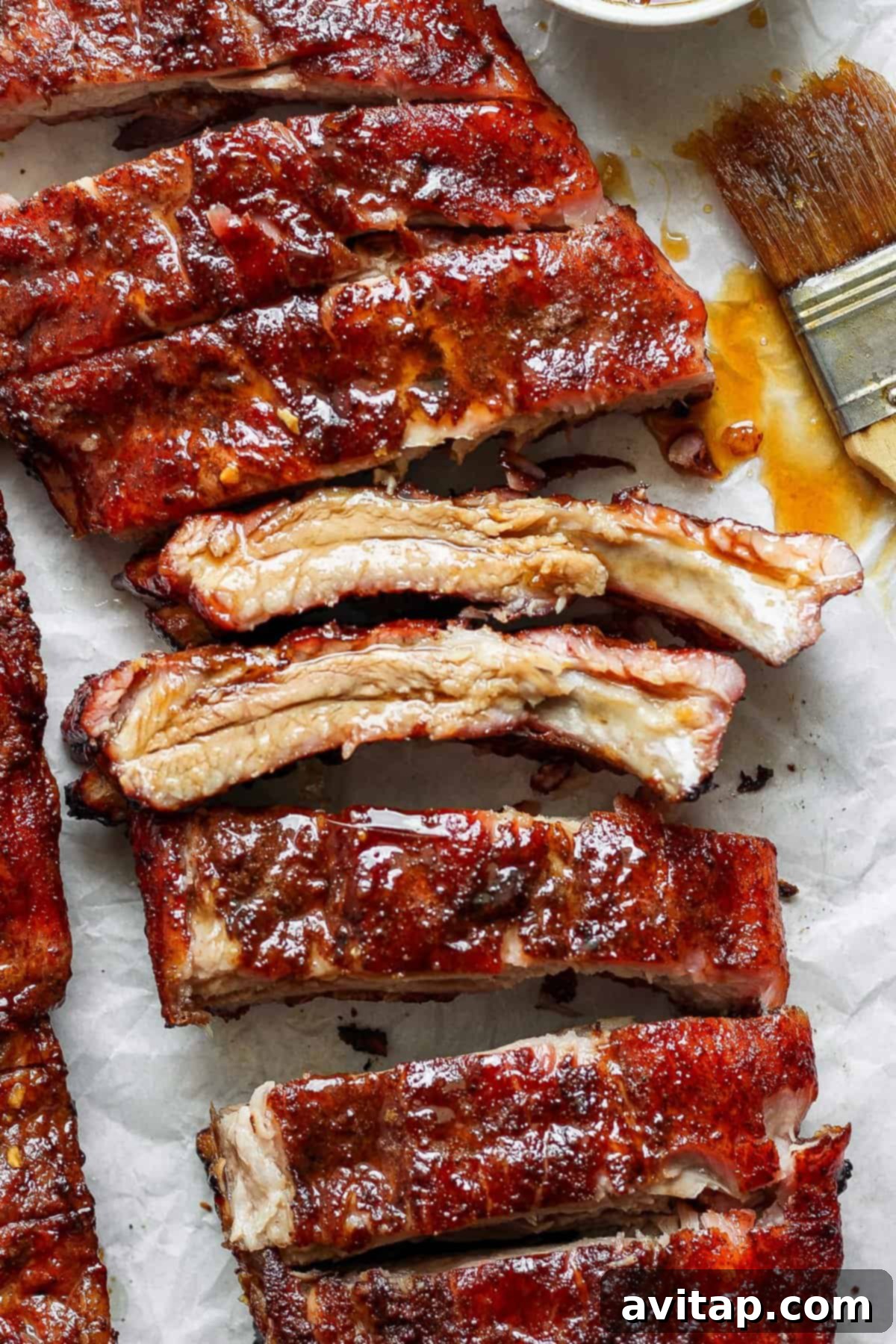 Close-up of cooked, glazed pork ribs on parchment paper with sauce and a basting brush beside them. The ribs are coated in a glossy, dark glaze.
