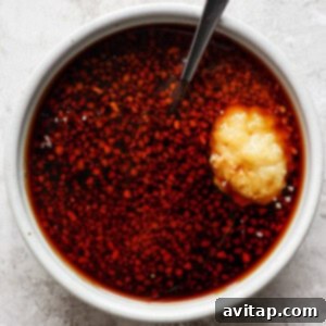 A white bowl filled with a reddish liquid and finely chopped ingredients, with a spoon and a dollop of minced garlic on top.