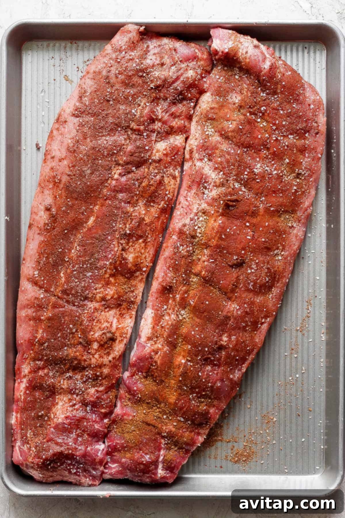 Two slabs of seasoned raw pork ribs are placed on a large rectangular baking sheet, ready for cooking.