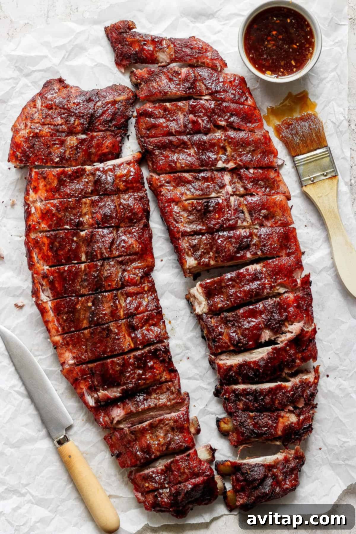 Two racks of barbecued ribs cut into portions, placed on parchment paper with a knife, a brush, and a bowl of sauce.