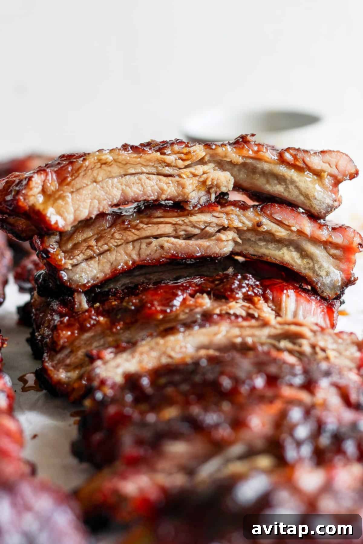 Close-up of a stack of glazed, grilled ribs, with the top piece cut to show a juicy, tender interior. The meat is richly caramelized with a glossy, charred exterior.