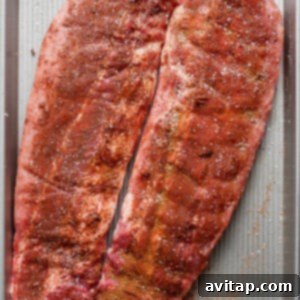 Two slabs of seasoned raw pork ribs are placed on a large rectangular baking sheet, ready for cooking.