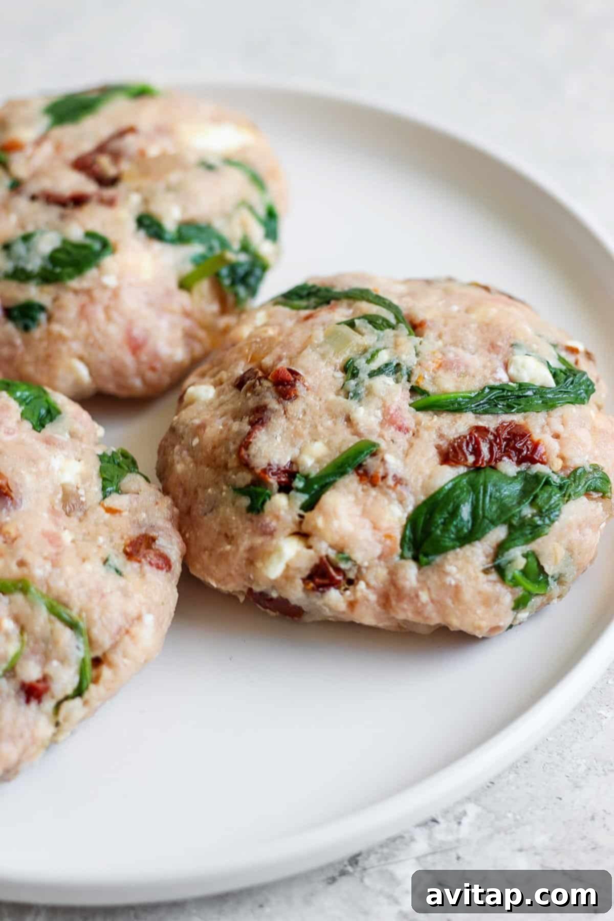 Hands gently mixing ground meat, spinach, sun-dried tomatoes, and feta cheese in a bowl for Mediterranean burgers.