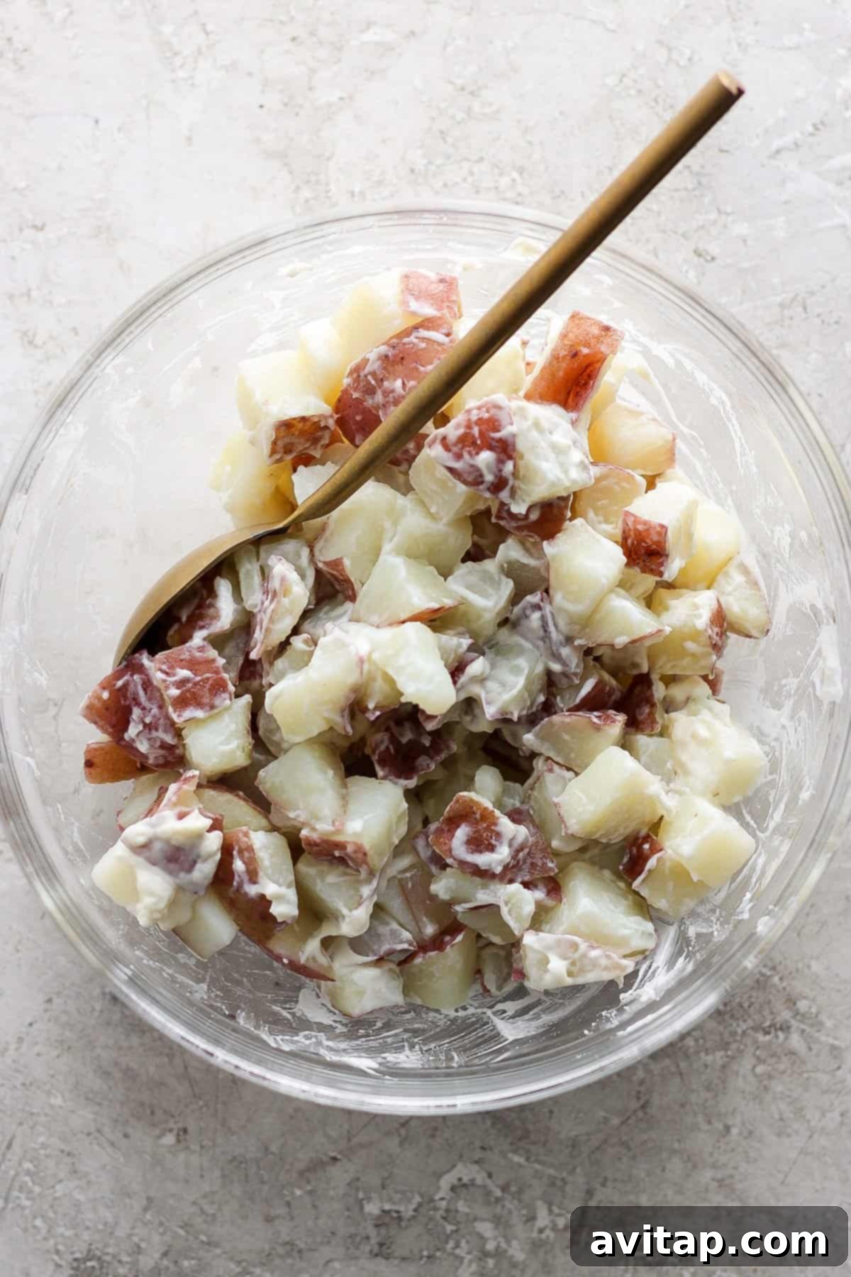 A bowl of creamy potato salad, with diced red potatoes, hints of fresh green herbs, and a light dressing, presented with a wooden spoon on a textured stone surface.
