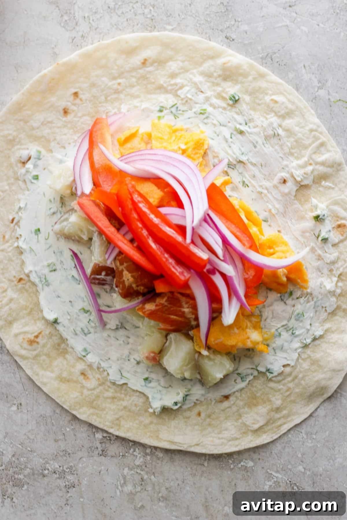 A large flour tortilla spread with a creamy herb cheese mixture, topped with vibrant slices of red bell pepper, red onion, and other fresh ingredients, ready for assembly.