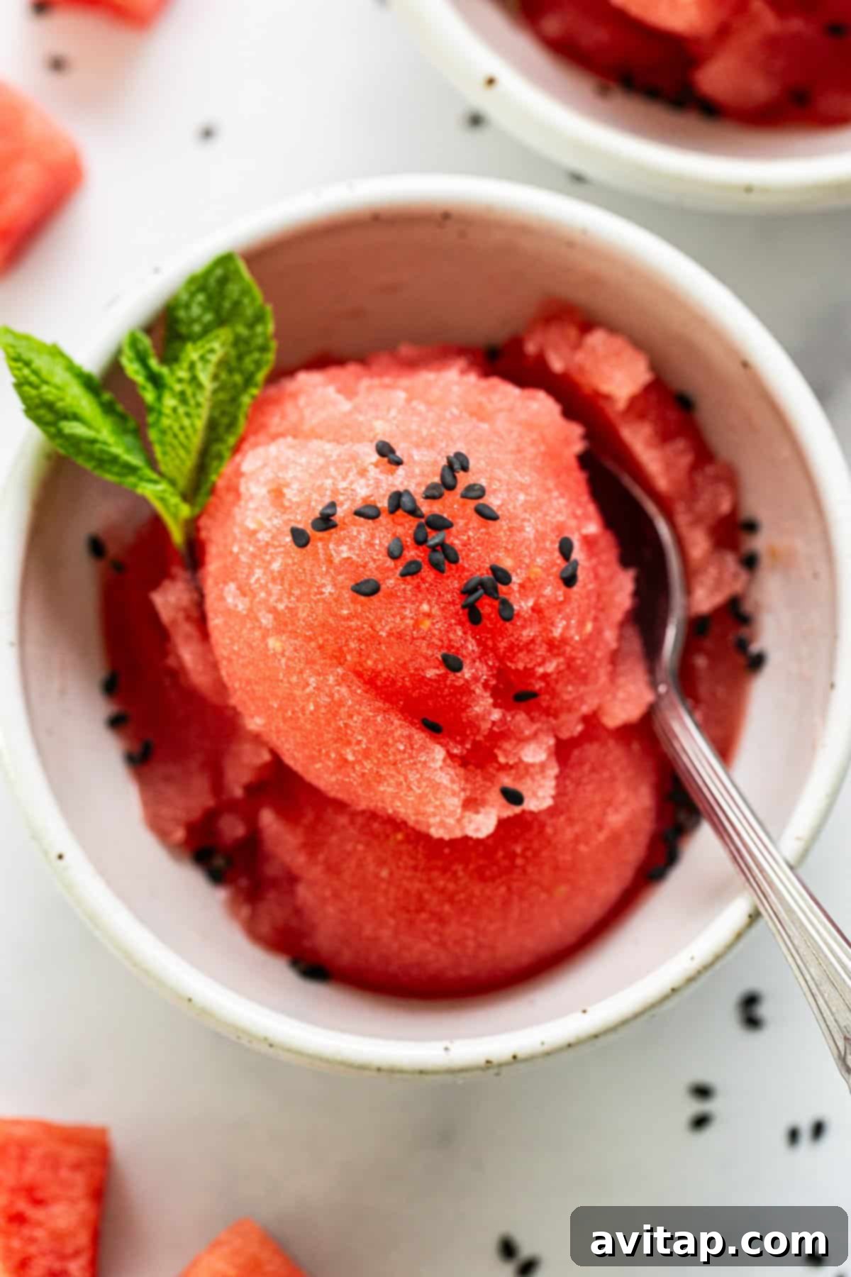 A vibrant bowl of homemade watermelon sorbet, beautifully garnished with fresh mint leaves and black sesame seeds, with a spoon resting beside it on a light, inviting surface, evoking a sense of cool refreshment.