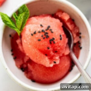 A bowl of watermelon sorbet garnished with mint leaves and black sesame seeds, accompanied by a spoon, on a light surface.