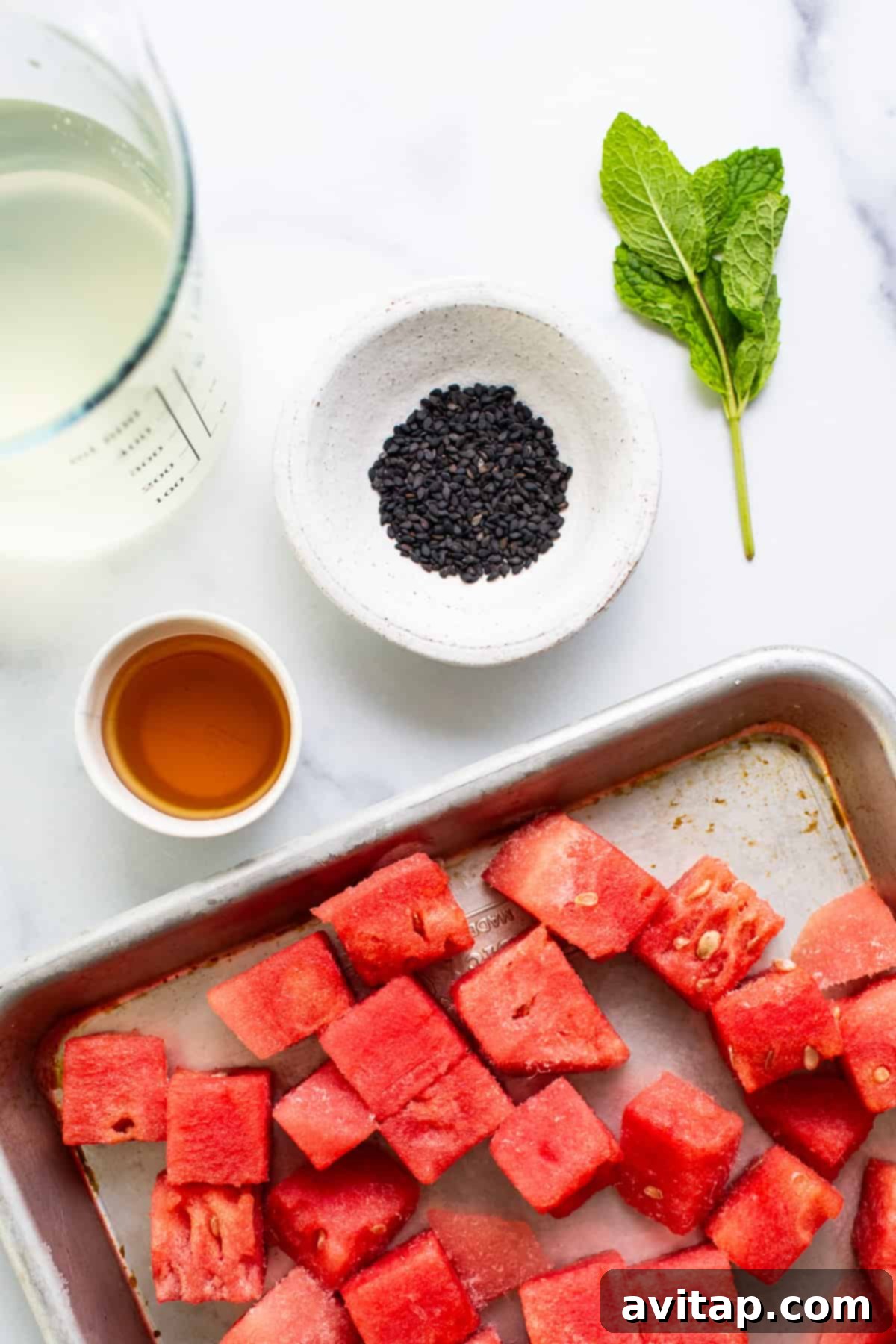 A beautifully composed flat lay featuring a tray of perfectly diced watermelon, a small elegant bowl filled with black sesame seeds, a fresh sprig of vibrant mint, a clear container of golden agave syrup, and a tall glass of pale coconut water, all arranged on a pristine white surface, highlighting the key ingredients for homemade watermelon sorbet.