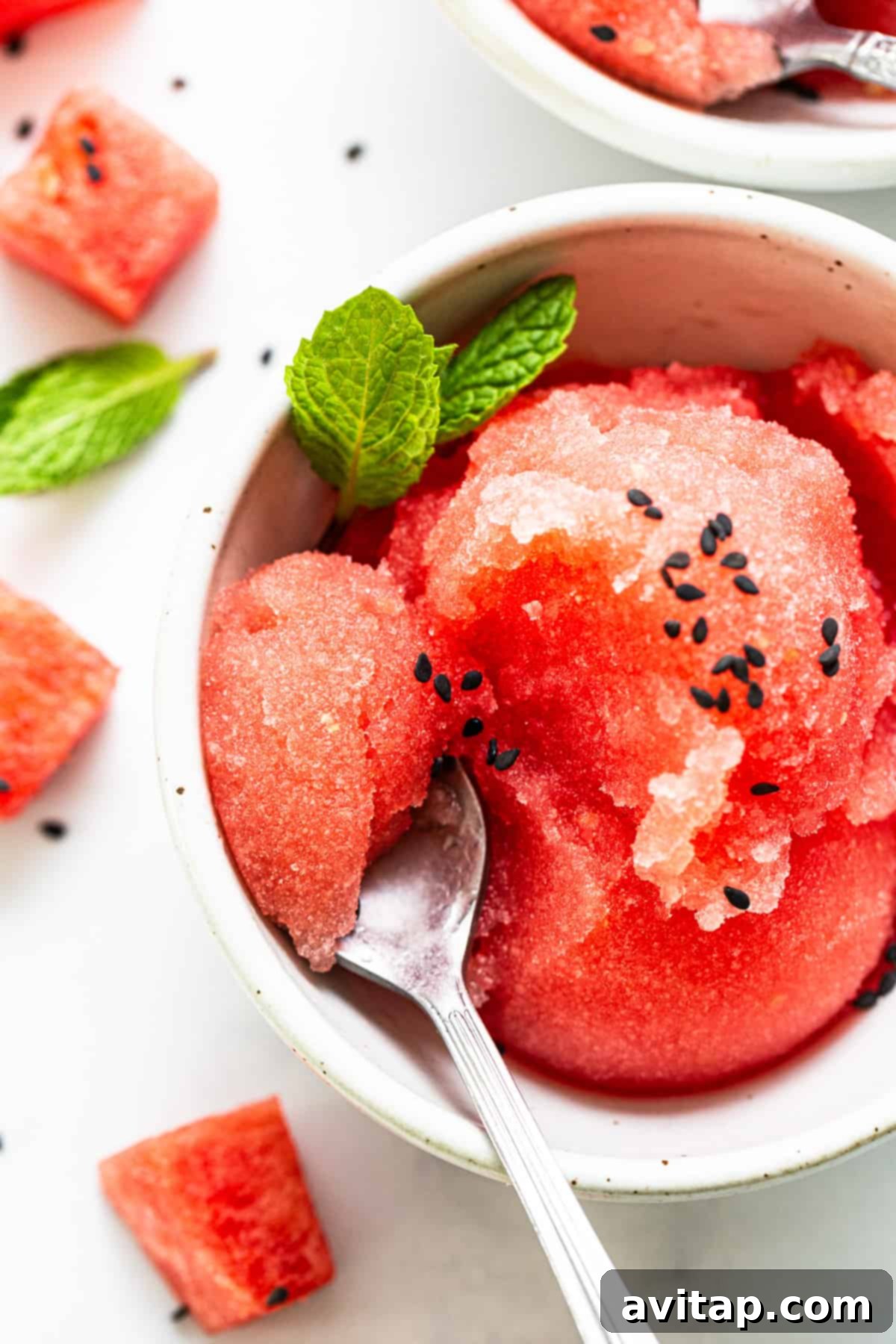 An enticing bowl of crimson watermelon sorbet, beautifully adorned with vibrant green mint leaves and tiny black sesame seeds, presented alongside a spoon and artfully scattered watermelon pieces on a clean white background, emphasizing its freshness and appeal.