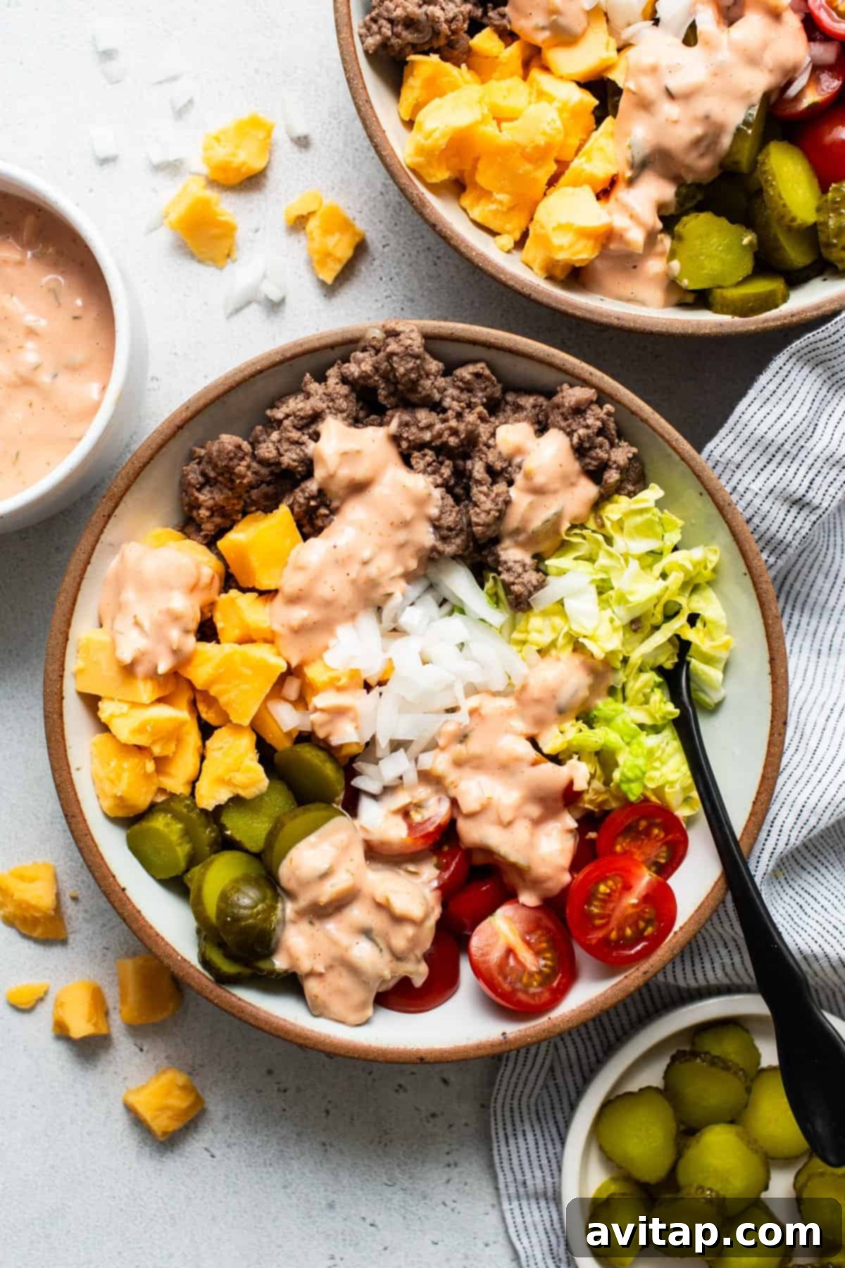 A colorful and hearty cheeseburger bowl featuring seasoned ground beef, crisp lettuce, juicy tomatoes, sliced pickles, and a generous drizzle of creamy burger sauce.