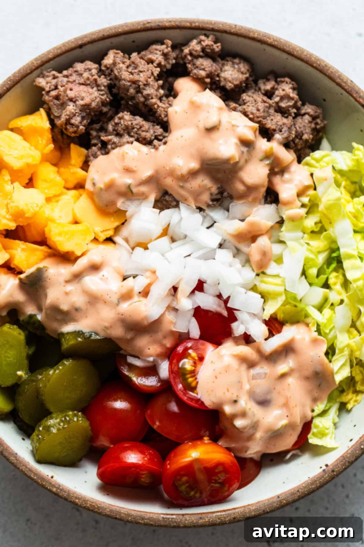 A perfectly arranged salad bowl showcasing distinct sections of seasoned ground beef, fresh chopped lettuce, juicy diced tomatoes, thinly sliced onions, and savory pickles, all lightly drizzled with a creamy dressing.