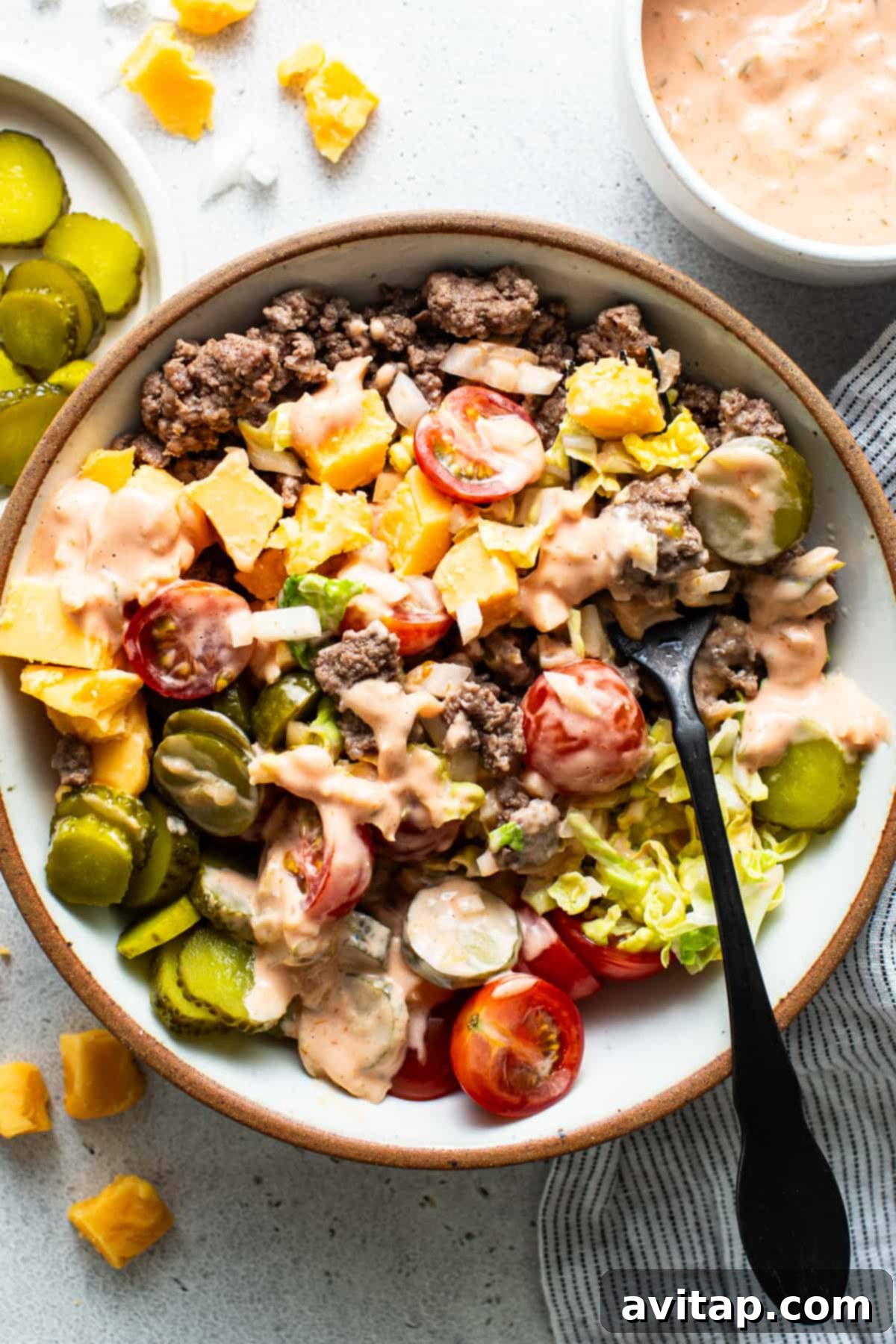 A delicious cheeseburger bowl featuring seasoned ground beef, fresh tomatoes, sliced pickles, shredded cheese, and crisp lettuce, all drizzled with a creamy dressing, with an extra side of sauce for dipping.