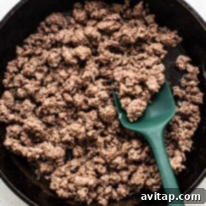 Cooked ground beef in a black cast iron skillet being stirred with a green spatula, showing browned meat and onions.