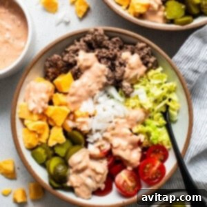 A beautifully assembled cheeseburger bowl with distinct sections of ground beef, fresh lettuce, tomatoes, pickles, and cheese, topped with creamy dressing.
