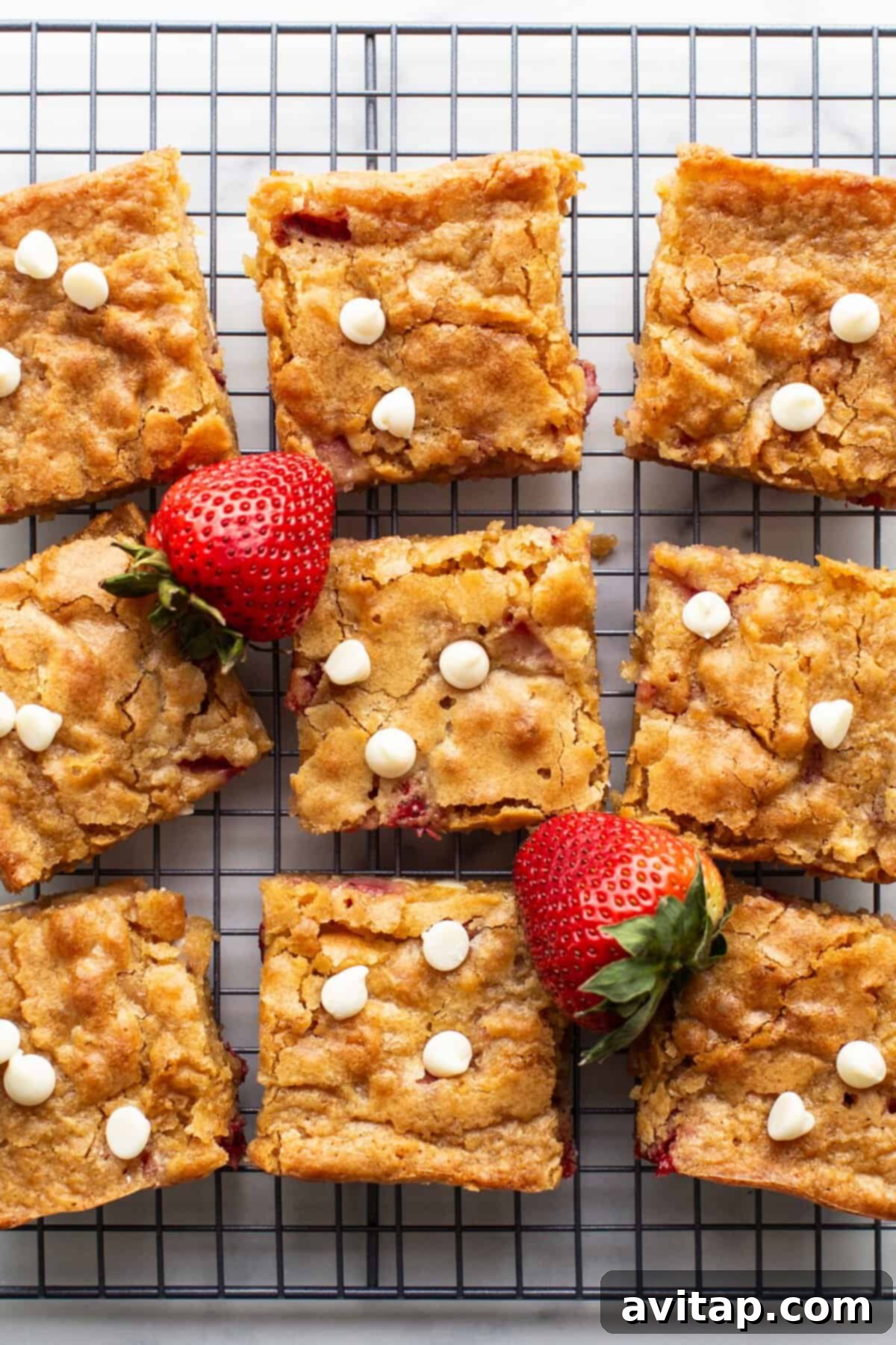 Nine square pieces of strawberry blondies with white chocolate chips and fresh strawberries rest on a cooling rack, showcasing their golden-brown edges and vibrant fruit pieces.