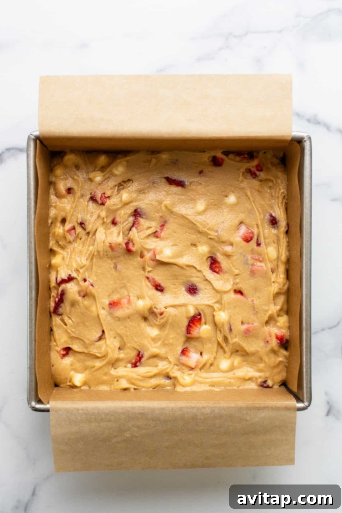 Square baking pan lined with parchment paper and filled with raw batter containing chunks of fresh strawberries and white chocolate chips, ready for the oven on a marble countertop.