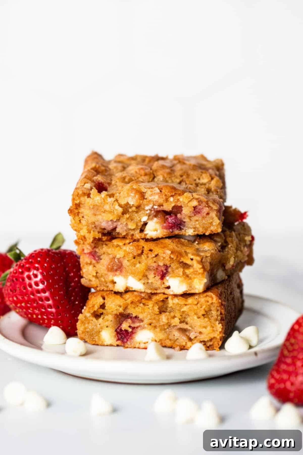 A plate of three stacked strawberry blondies, generously studded with white chocolate chips and pieces of strawberry, artfully arranged with fresh strawberries and loose white chocolate chips.