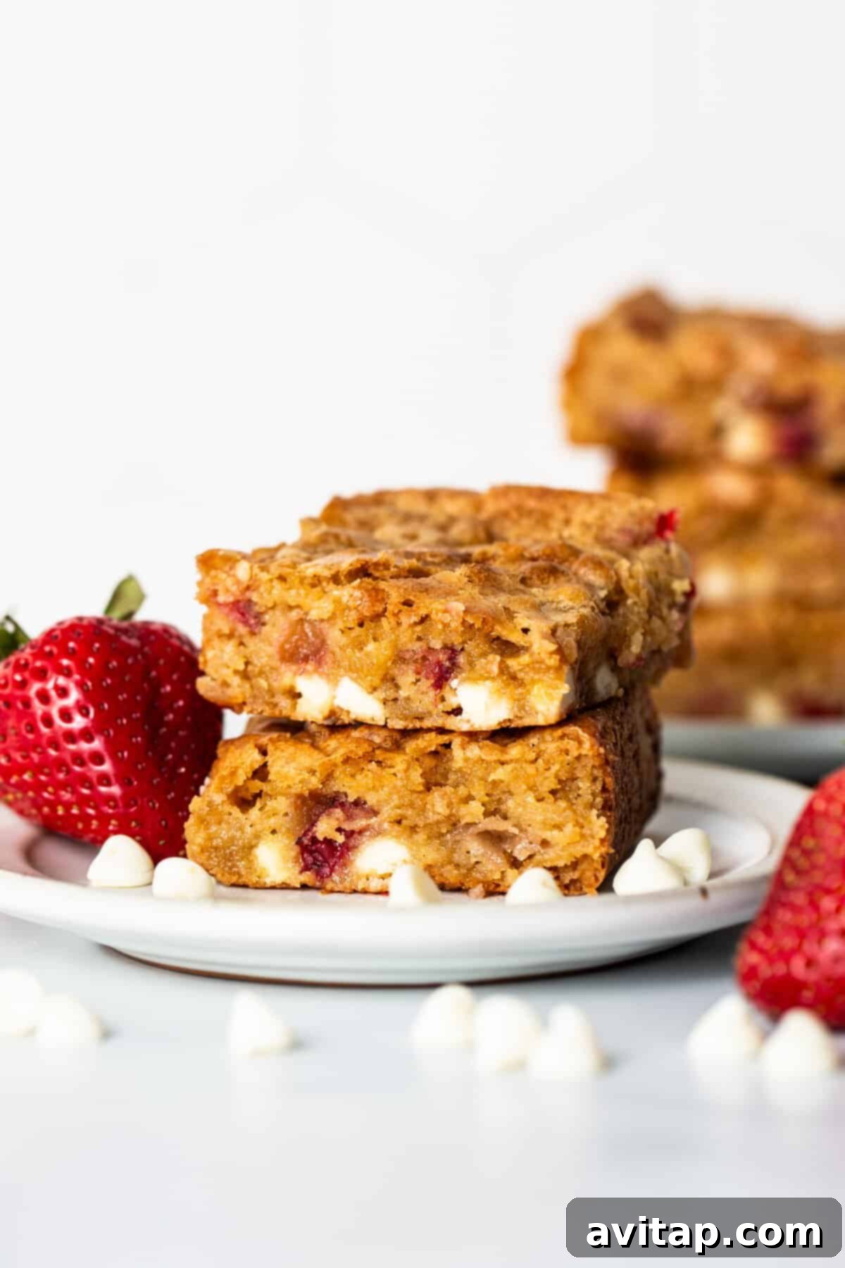 Close-up of two stacked strawberry blondies on a small plate, revealing visible strawberries and white chocolate chips, accompanied by whole strawberries and scattered white chocolate chips, highlighting their appealing texture.