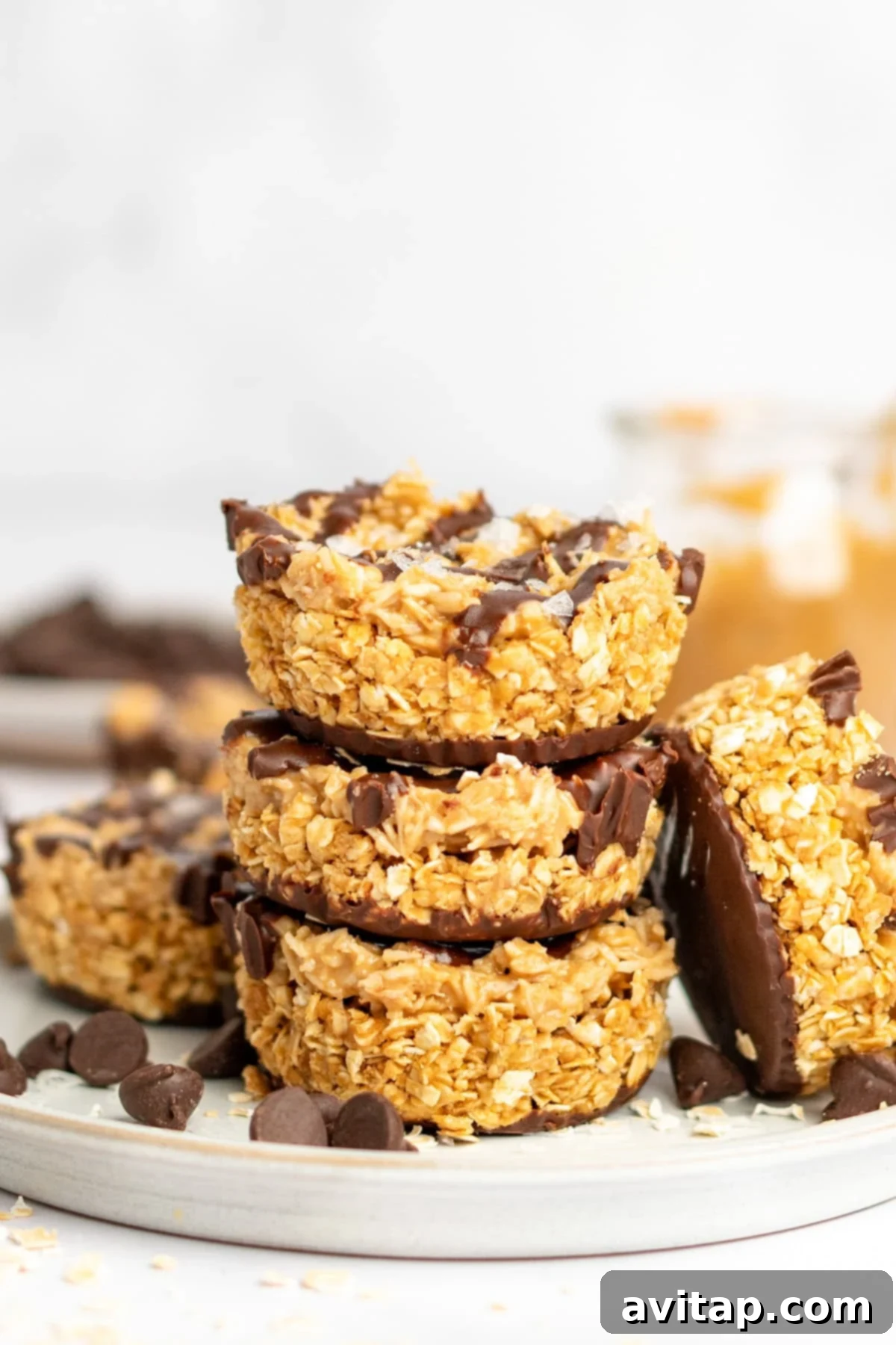 A stack of four oat and chocolate dessert bars on a plate with chocolate chips scattered around. A jar with peanut butter is visible in the background.