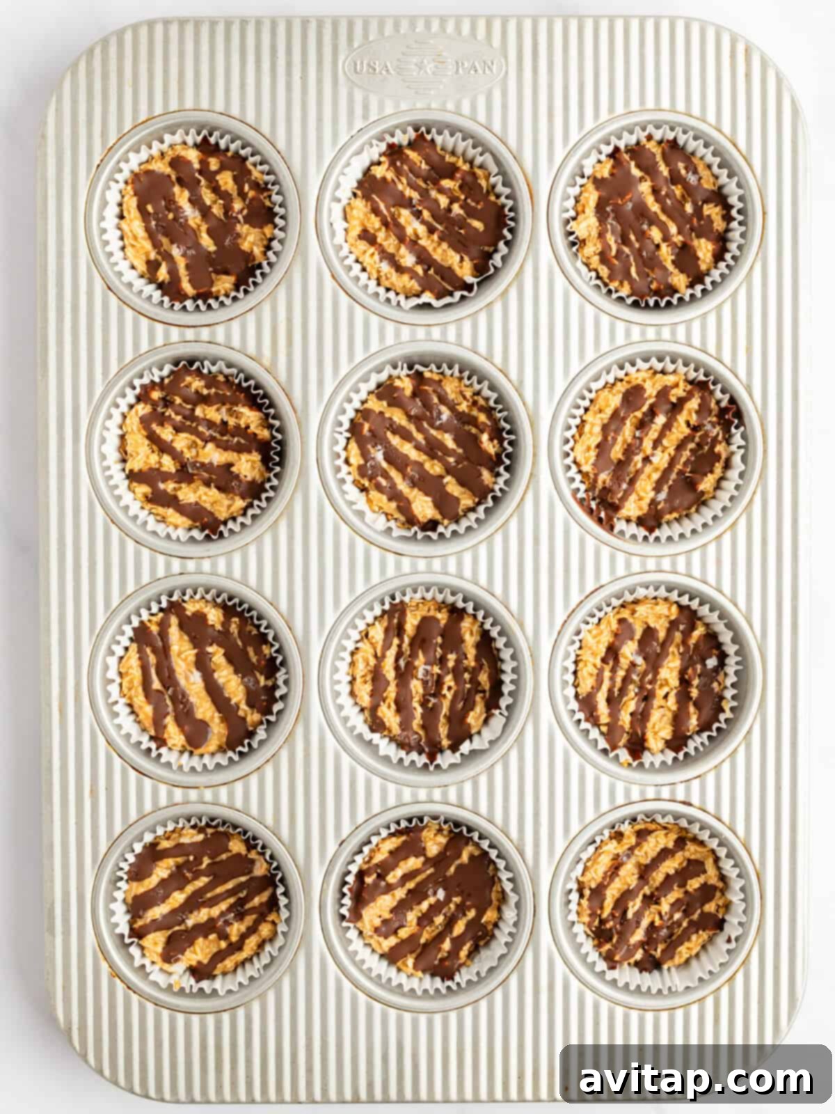A muffin tin filled with 12 chocolate-drizzled oatmeal cookies in white paper liners, ready to be set.