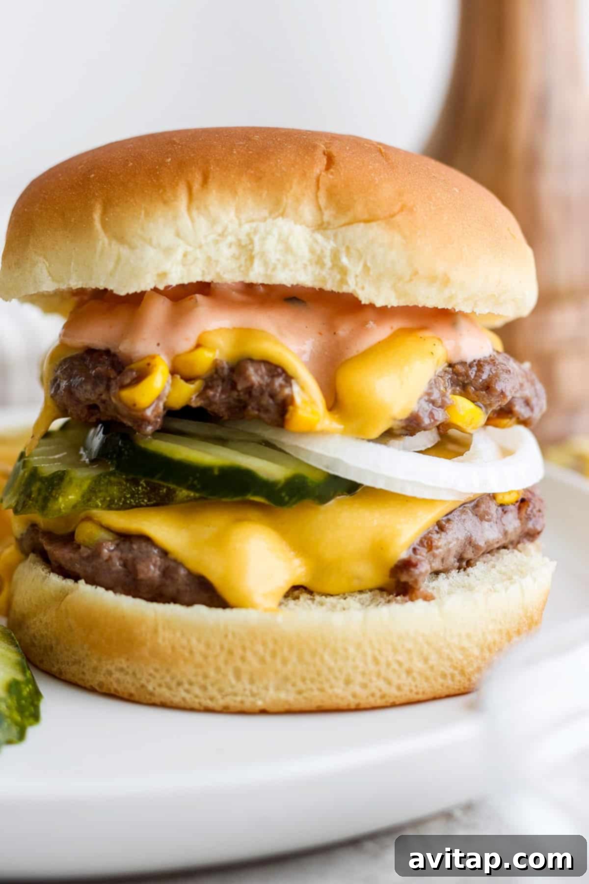 Sweet Corn Medley Smash Burger 6 A double cheeseburger with pickles, onions, and sauce on a white plate, showcasing its height and layers.