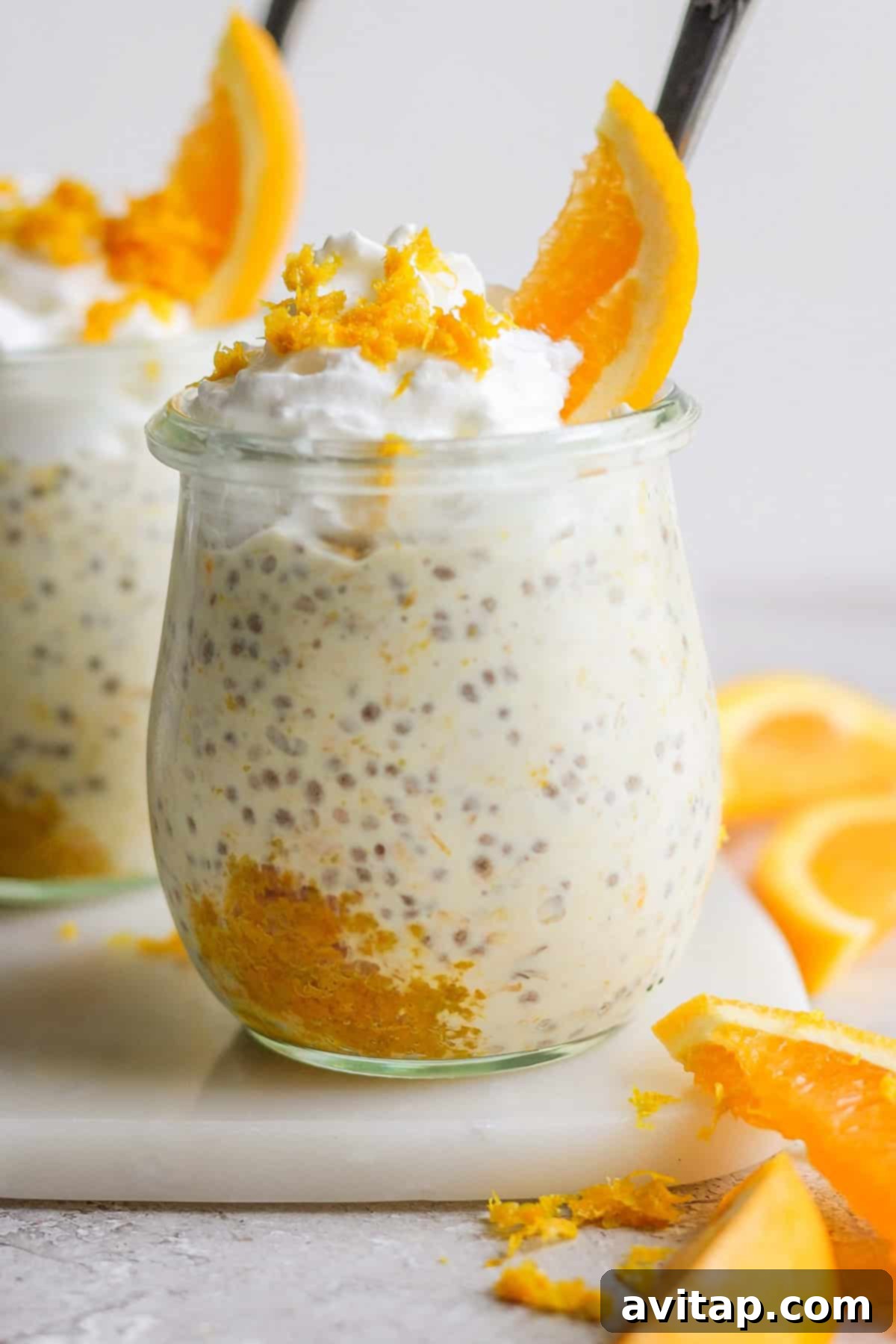 Sunshine Creamsicle Overnight Oats 2 A glass jar filled with chia seed pudding topped with whipped cream, orange zest, and an orange slice, giving it a delightful creamsicle overnight oats feel, accompanied by pieces of orange on a white surface.