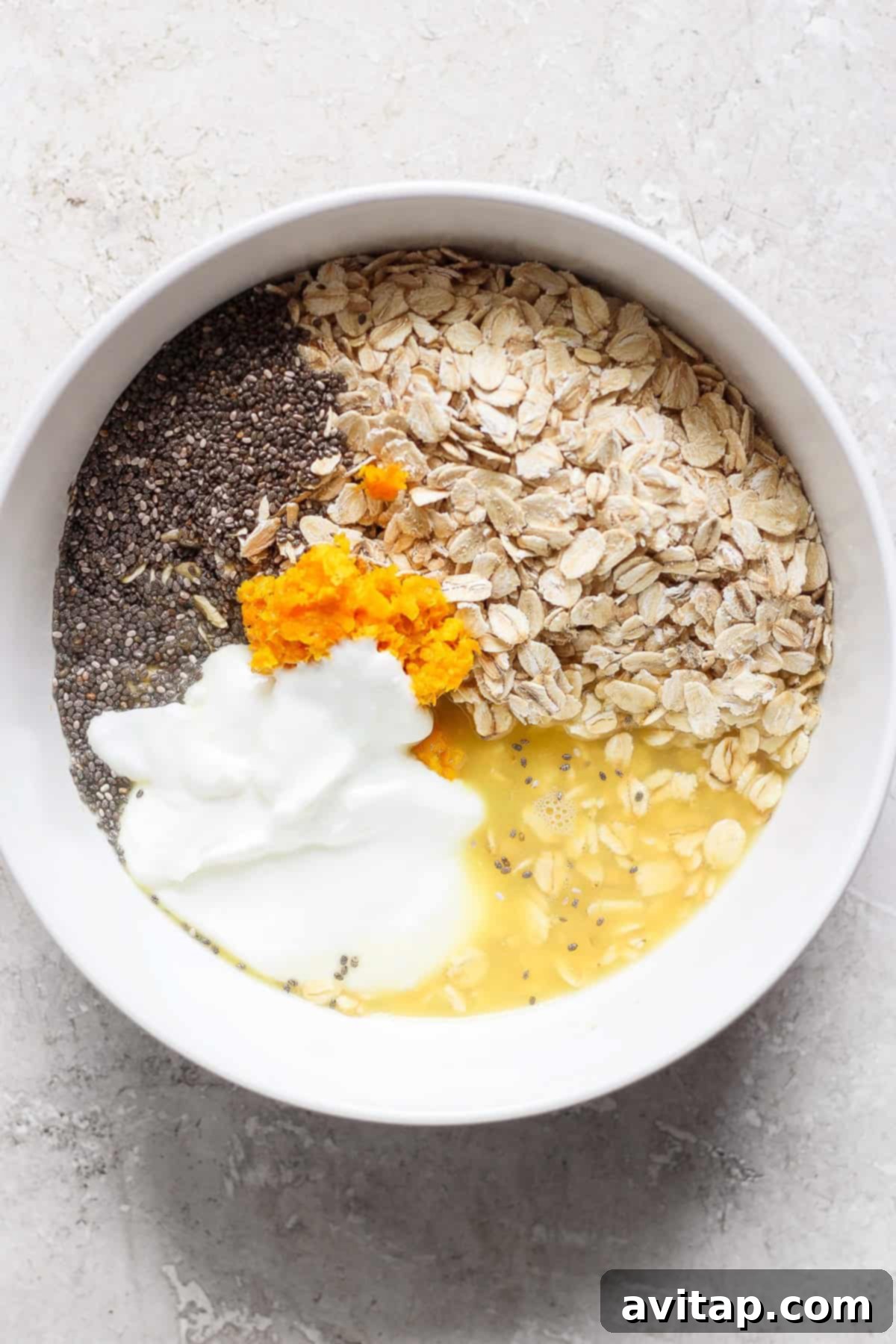 Sunshine Creamsicle Overnight Oats 3 A white bowl contains oats, chia seeds, orange zest, yogurt, and liquid—likely milk or water—on a light-colored surface, creating a creamsicle overnight oats sensation.