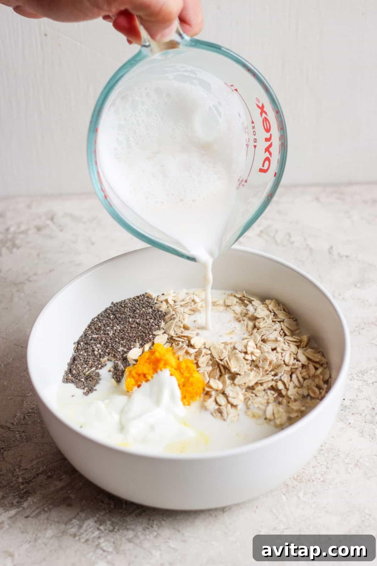 Sunshine Creamsicle Overnight Oats 4 A hand pours milk from a measuring cup into a white bowl containing oats, chia seeds, orange zest, and yogurt, creating the perfect base for creamsicle overnight oats.