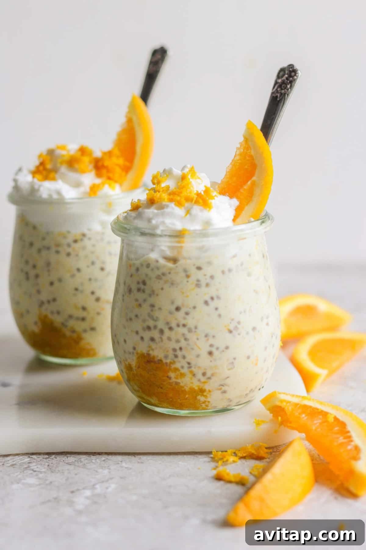 Sunshine Creamsicle Overnight Oats 6 Two glass jars filled with creamy creamsicle overnight oats, topped with whipped cream and orange slices, arranged on a white surface with additional orange wedges nearby.
