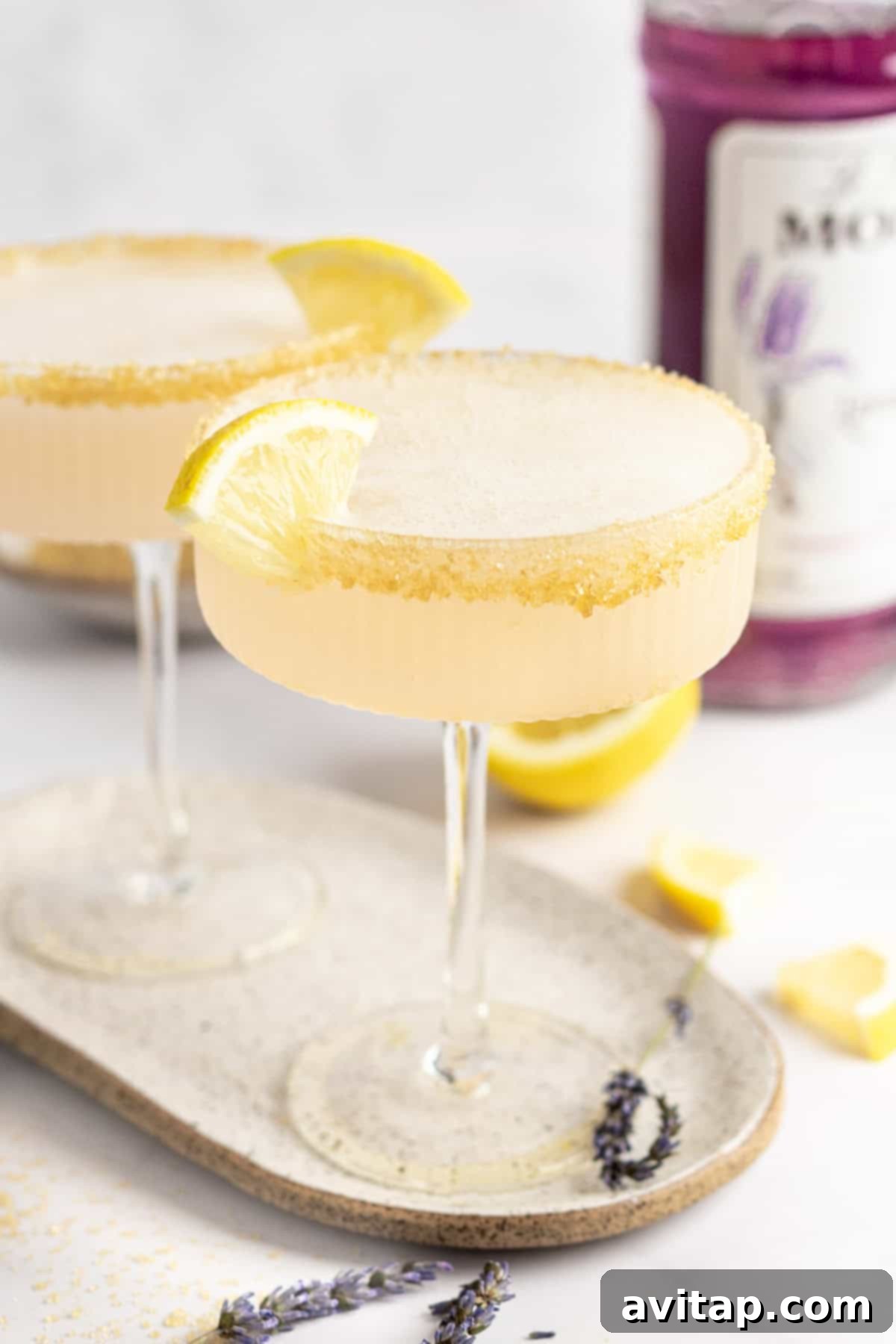 Two coupe glasses with yellow cocktails garnished with lemon slices and sugar rims on a beige tray. Purple-hued bottle in the background. Lavender sprigs and lemon pieces scattered around.