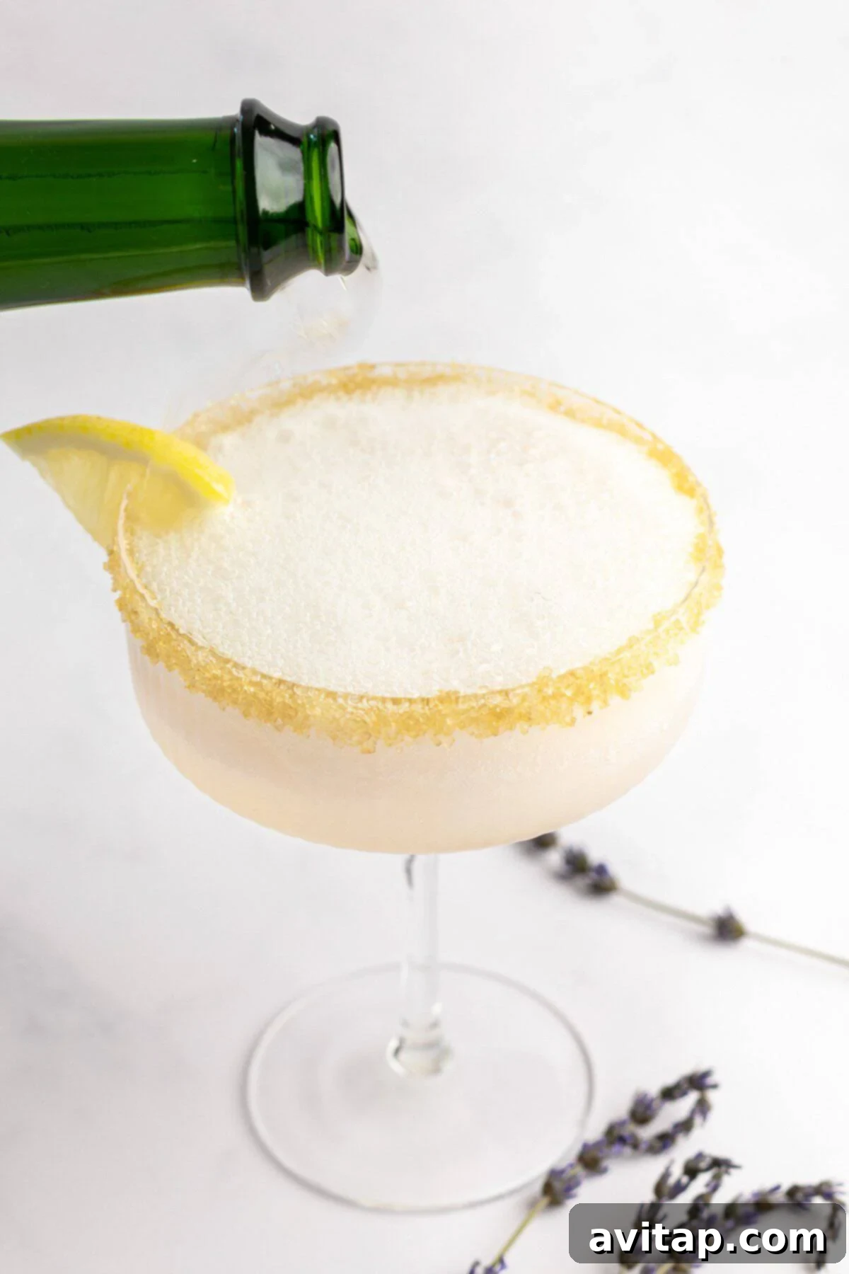 A green bottle pours liquid into a frothy glass with a sugared rim, garnished with a lemon slice. Lavender sprigs lie next to the glass on a white background.