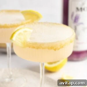 Two cocktails with frothy tops in coup glasses, each garnished with a lemon wedge and a sugar-rimmed edge. A bottle of purple liquid is partially visible in the background.