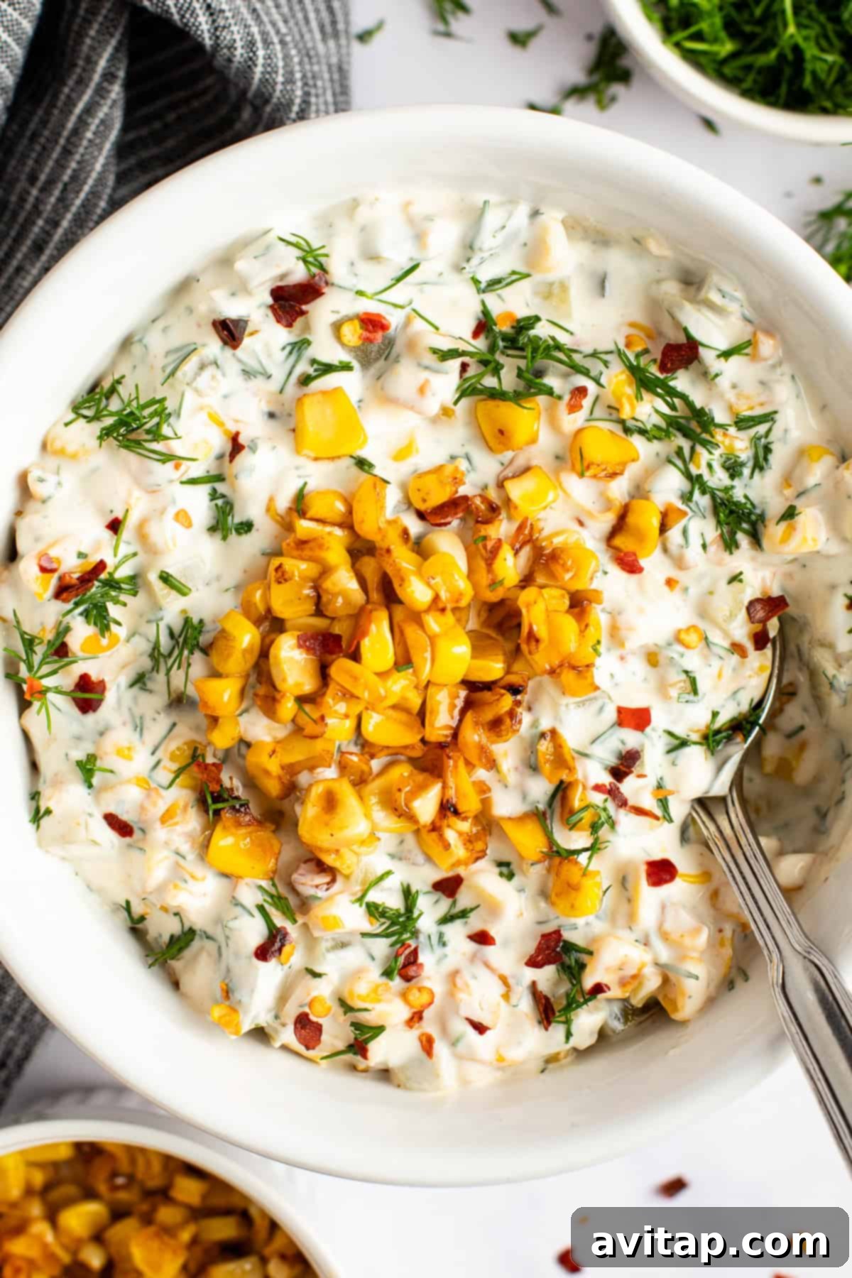 A bowl of creamy corn dip garnished with grilled corn, fresh herbs, and red pepper flakes, with a spoon resting inside the bowl. Perfect for a party appetizer.
