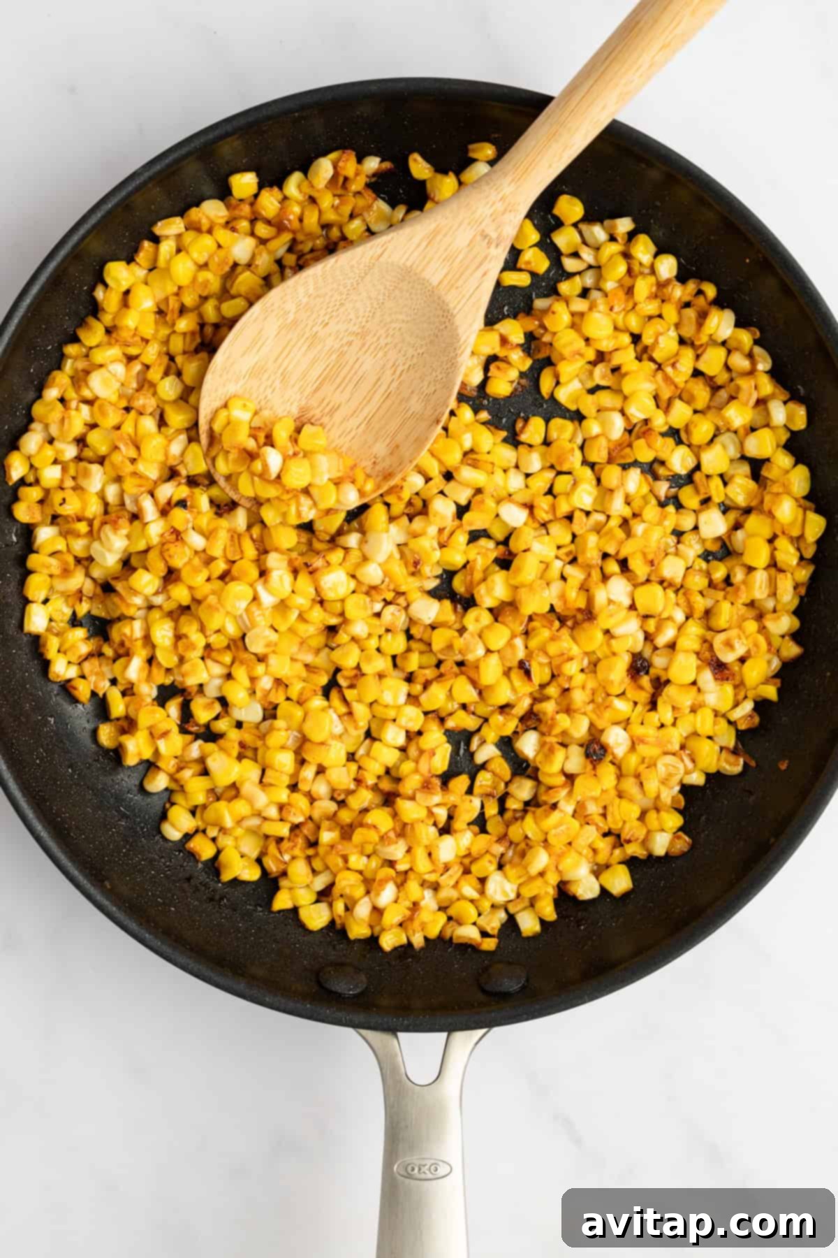 Sautéed corn kernels in a black frying pan with a wooden spoon on a white countertop, highlighting the browning process for a creamy corn dip.
