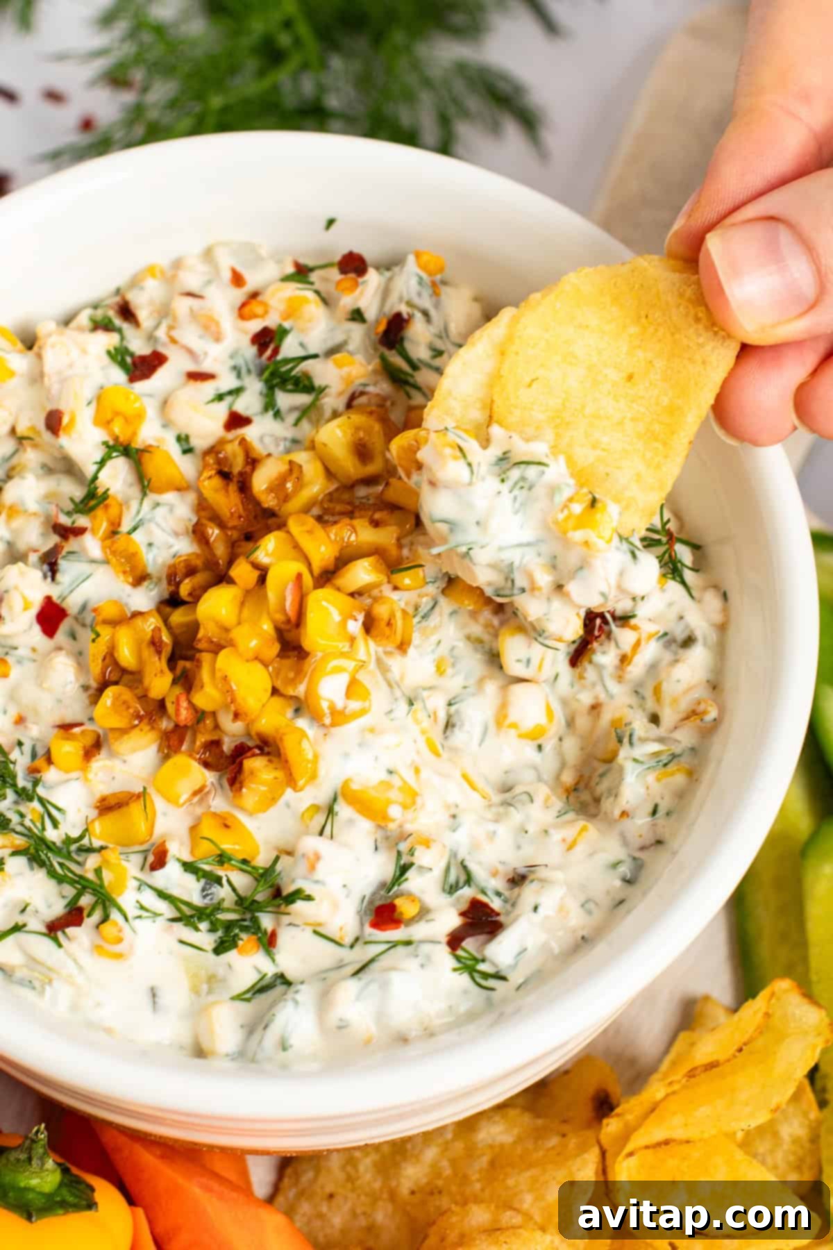 A hand dips a chip into a bowl of creamy dip topped with corn kernels and herbs, surrounded by chips and vegetable slices, perfect for sharing.