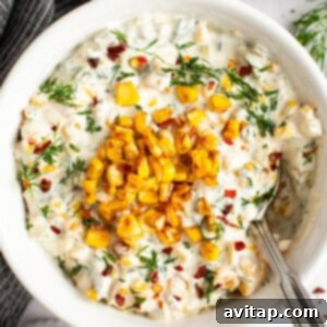 A bowl of creamy dip topped with roasted corn, chopped herbs, and red pepper flakes, with a spoon on the side.