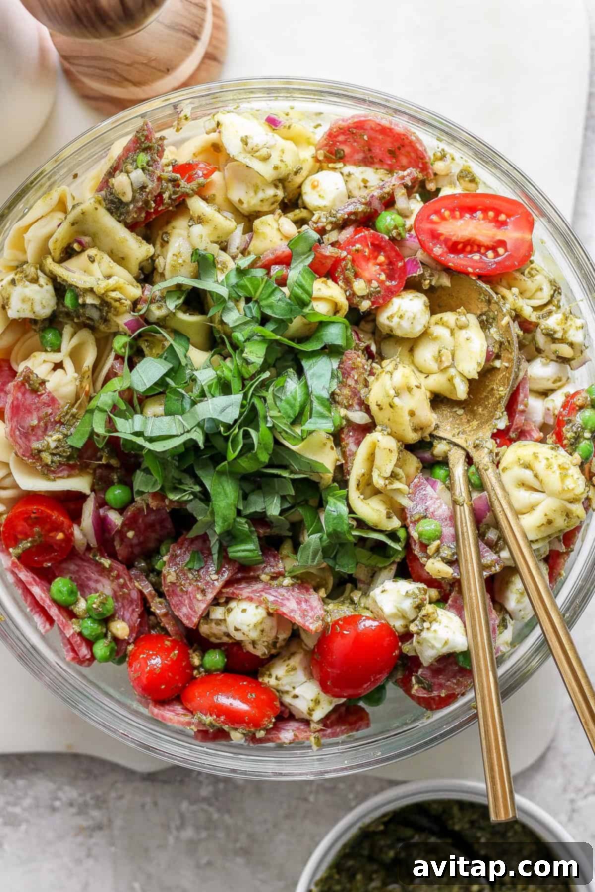 Vibrant Pesto Tortellini Medley 2 A bowl of tortellini pasta salad with cherry tomatoes, mozzarella, peas, salami, and greens, served with pesto and two spoons.