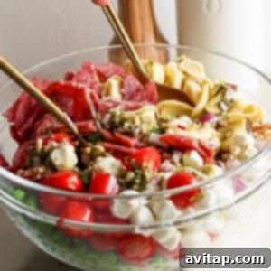A glass bowl containing ingredients like cherry tomatoes, small mozzarella balls, salami slices, and pasta being mixed with spoons. A bottle and wooden salt grinder are in the background.