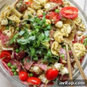 A bowl of tortellini pasta salad with cherry tomatoes, mozzarella, peas, salami, and greens, served with pesto and two spoons.