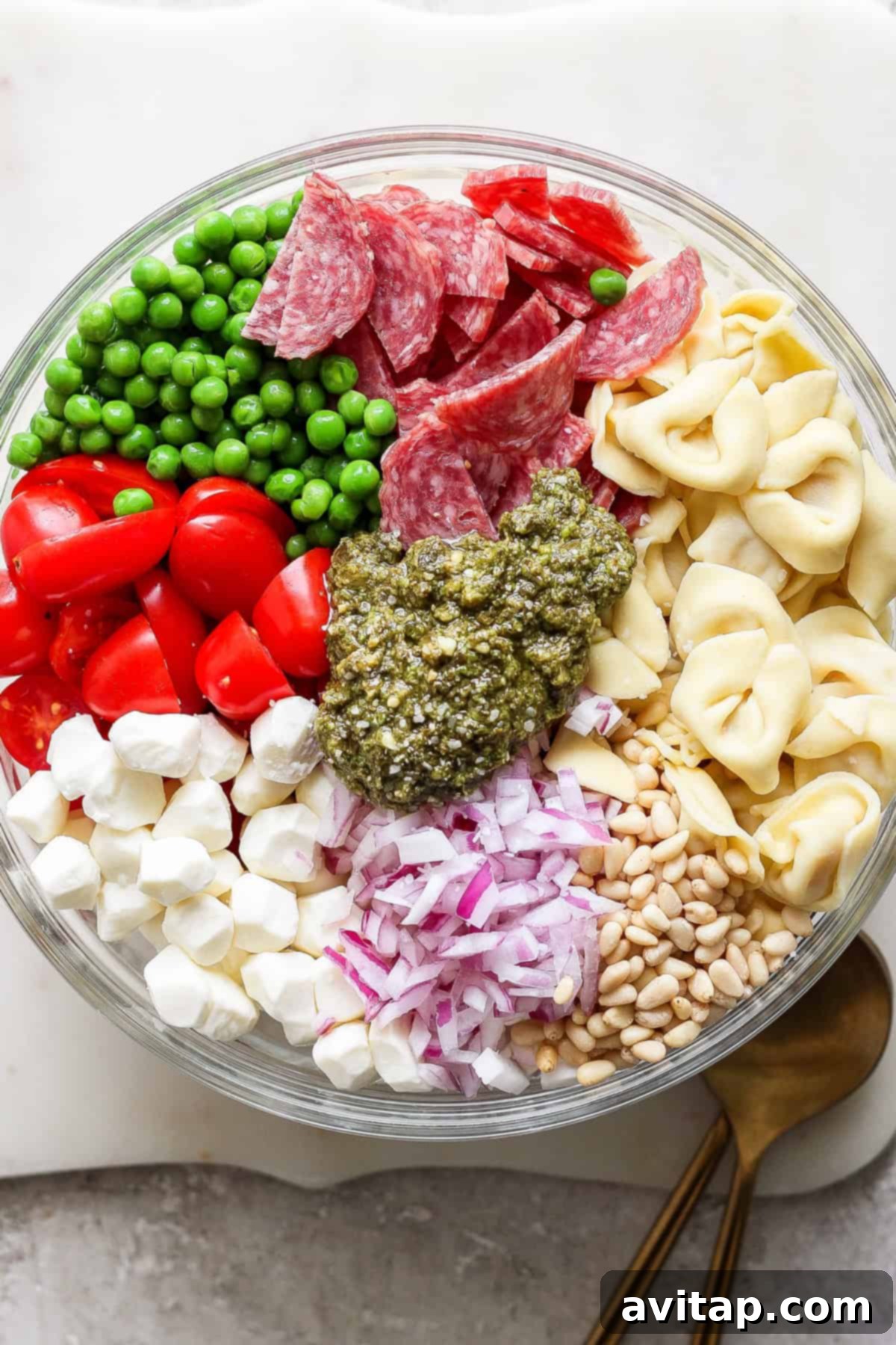 Vibrant Pesto Tortellini Medley 3 A glass bowl filled with peas, salami slices, cherry tomatoes, mozzarella balls, diced red onions, pine nuts, tortellini, and a dollop of pesto in the center. Two wooden utensils lie beside the bowl.