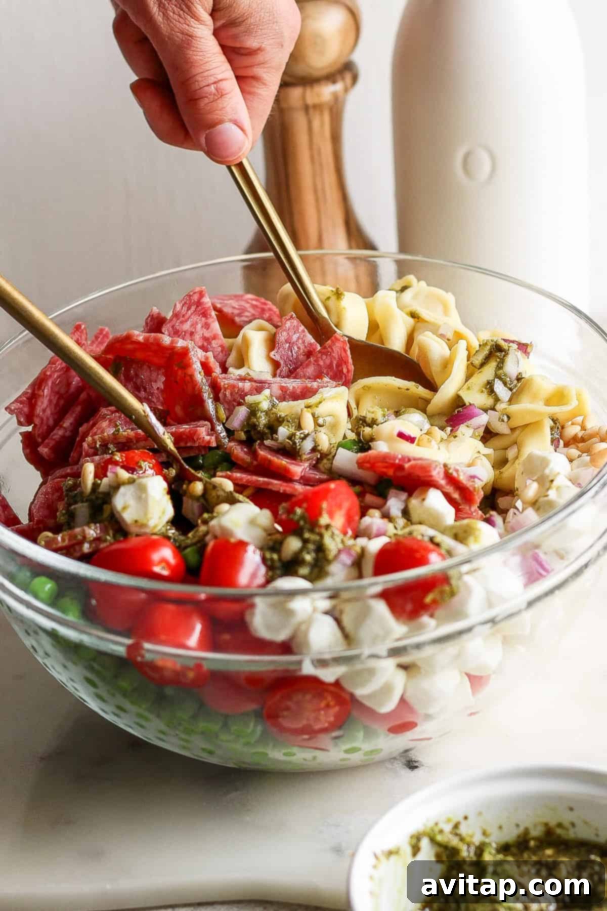 Vibrant Pesto Tortellini Medley 4 A glass bowl containing ingredients like cherry tomatoes, small mozzarella balls, salami slices, and pasta being mixed with spoons. A bottle and wooden salt grinder are in the background.