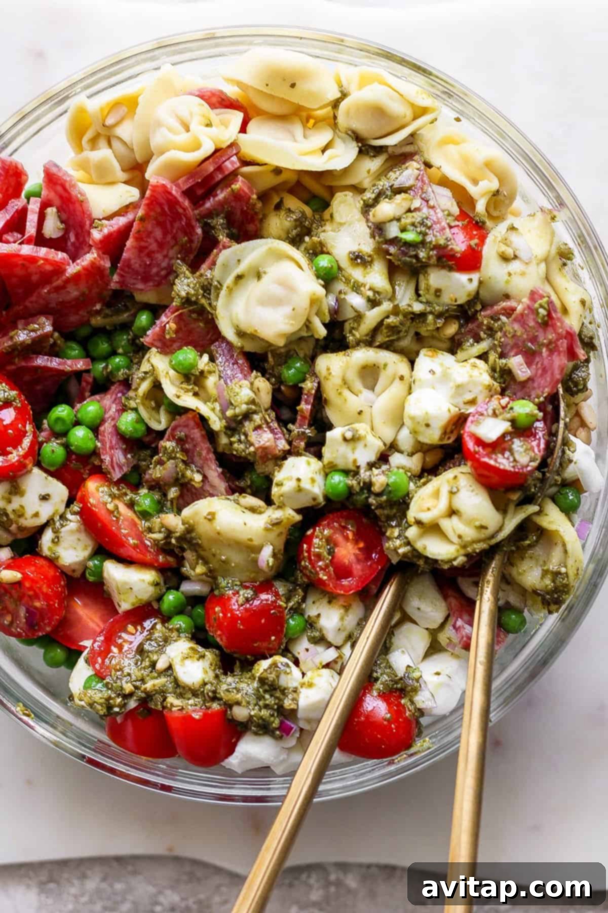 Vibrant Pesto Tortellini Medley 6 A bowl of pasta salad with tortellini, cherry tomatoes, peas, sliced meat, and pesto sauce, with two wooden utensils placed inside.