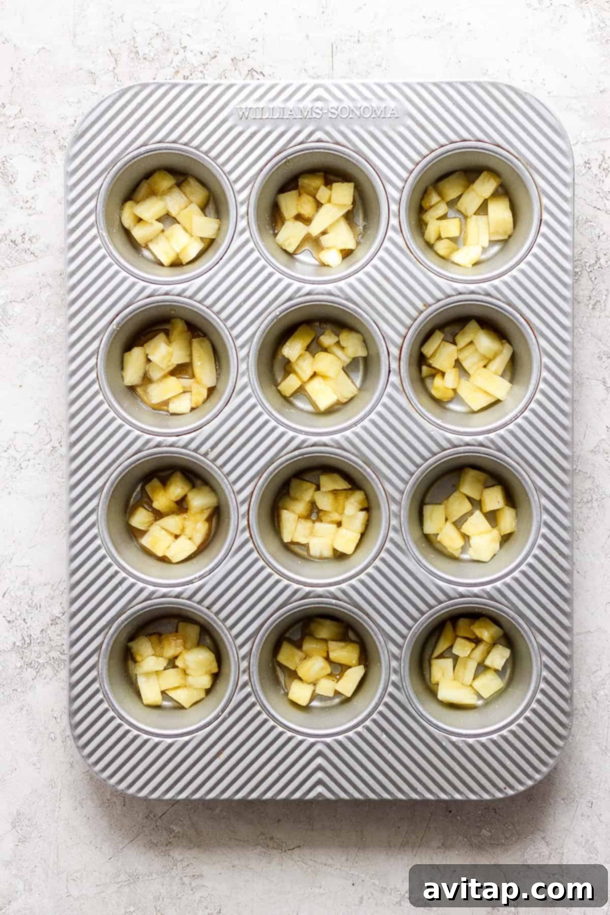 A muffin tin containing evenly chopped apples in each compartment, set on a light textured surface.