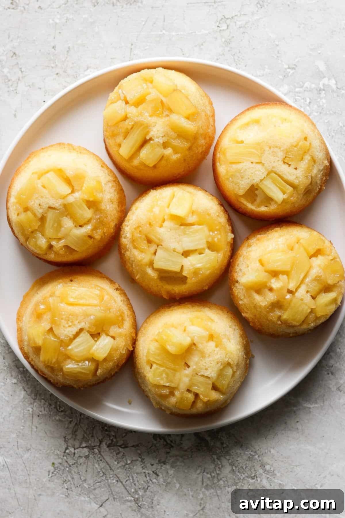 Plate of pineapple upside-down mini cakes topped with pineapple chunks on a light textured surface.