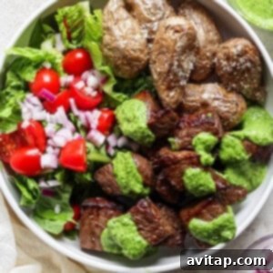 Effortless Air Fryer Steak Bites 12 A bowl of grilled steak chunks with green sauce, roasted potatoes, and a side salad of lettuce, tomatoes, and chopped red onions.