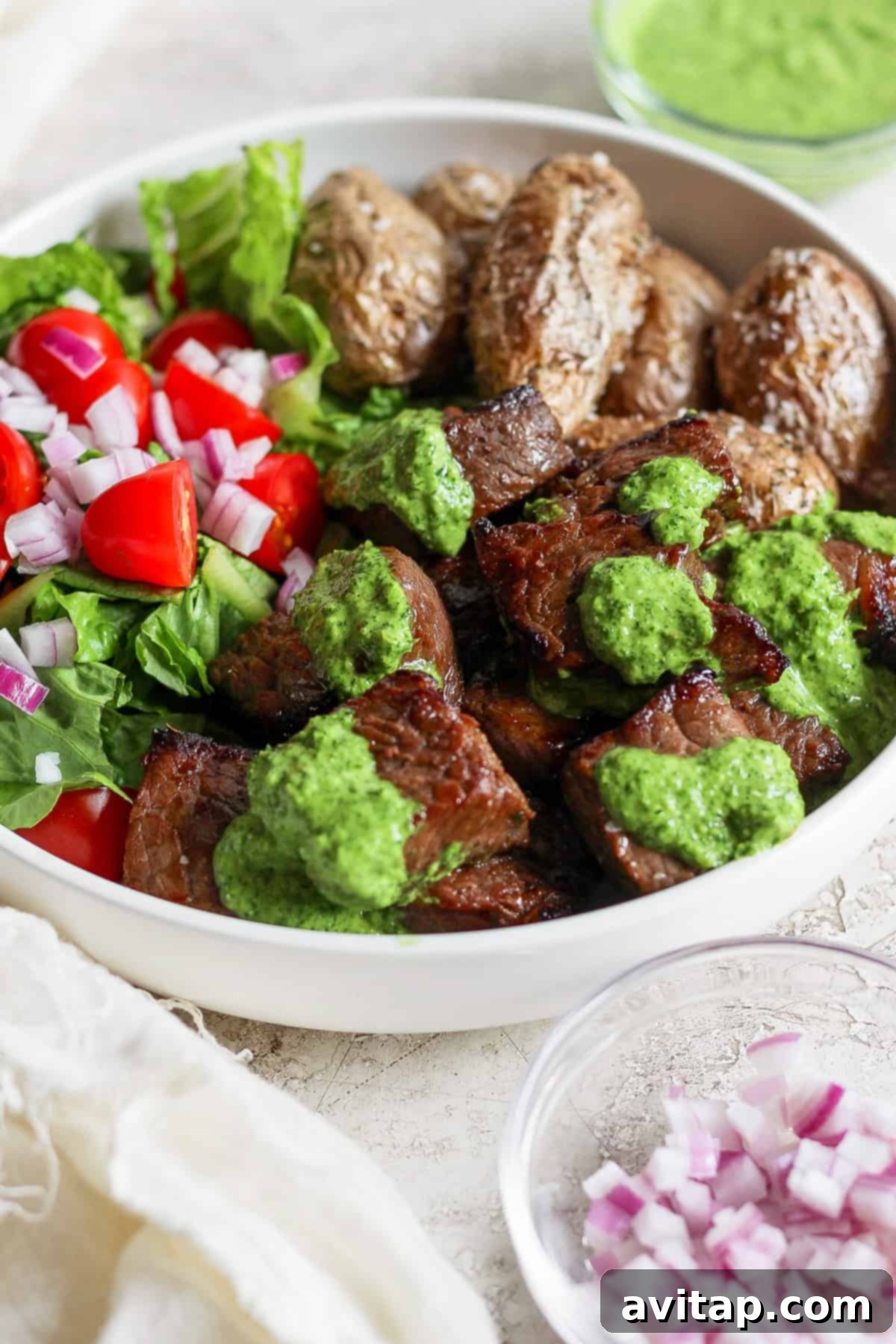 Effortless Air Fryer Steak Bites 7 A visually appealing bowl featuring grilled steak, richly covered with a vibrant green chimi-churri sauce, alongside perfectly roasted potatoes and a fresh salad composed of crisp lettuce, bright cherry tomatoes, and finely chopped red onions. A small bowl of extra chopped red onions sits beside it.